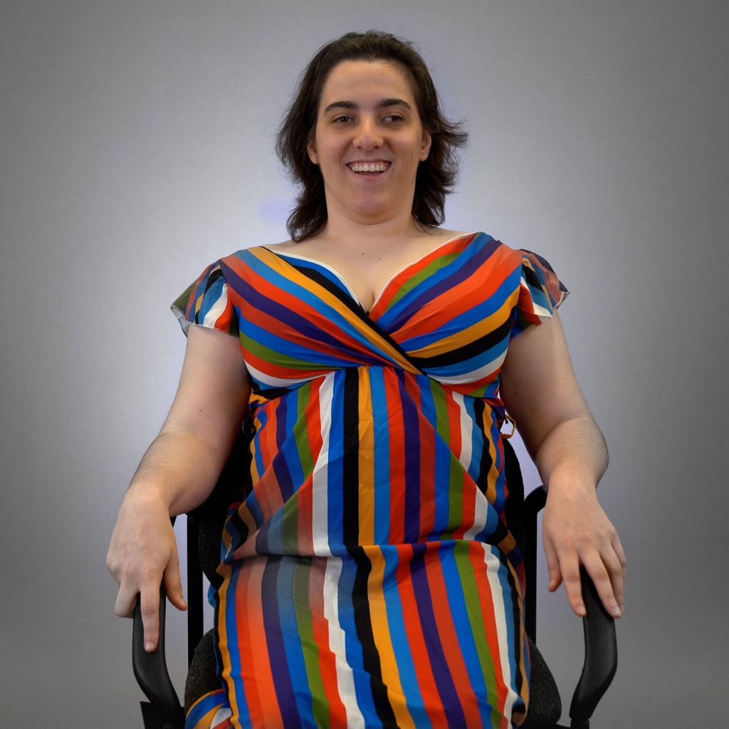 A person with medium-length dark hair smiles while seated against a plain grey background. They are wearing a colourful dress with vertical and diagonal stripes in red, blue, green, orange, black, and white.
