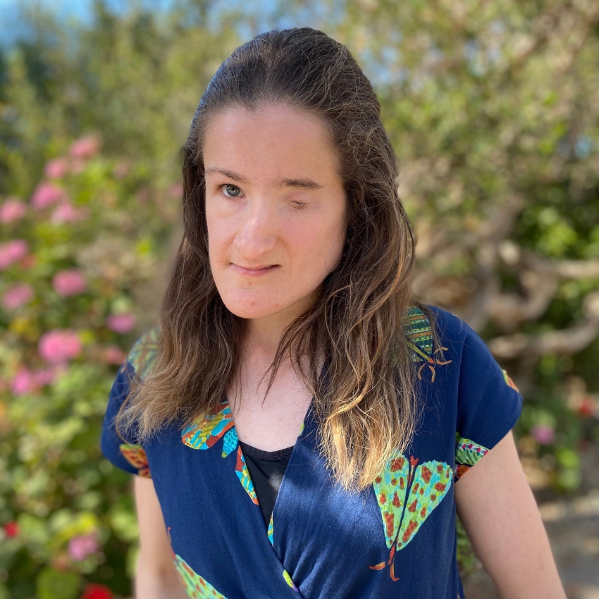 A woman with long brown hair stands outdoors in front of a blurred background of greenery and pink flowers. She is wearing a navy blue jumpsuit with a colourful print and looks calmly toward the camera.