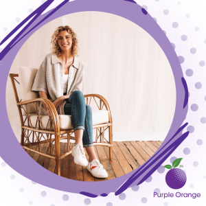 a white to light purple gradient with darker purple dots in the background. The Purple Orange logo in the bottom right corner. A grey purple misshapen circle with dark purple lines borders a photo of Lauren, who sits in a wooden chair wearing blue jeans and light coloured tops. Lauren smiles at the camera and has shoulder length curly hair
