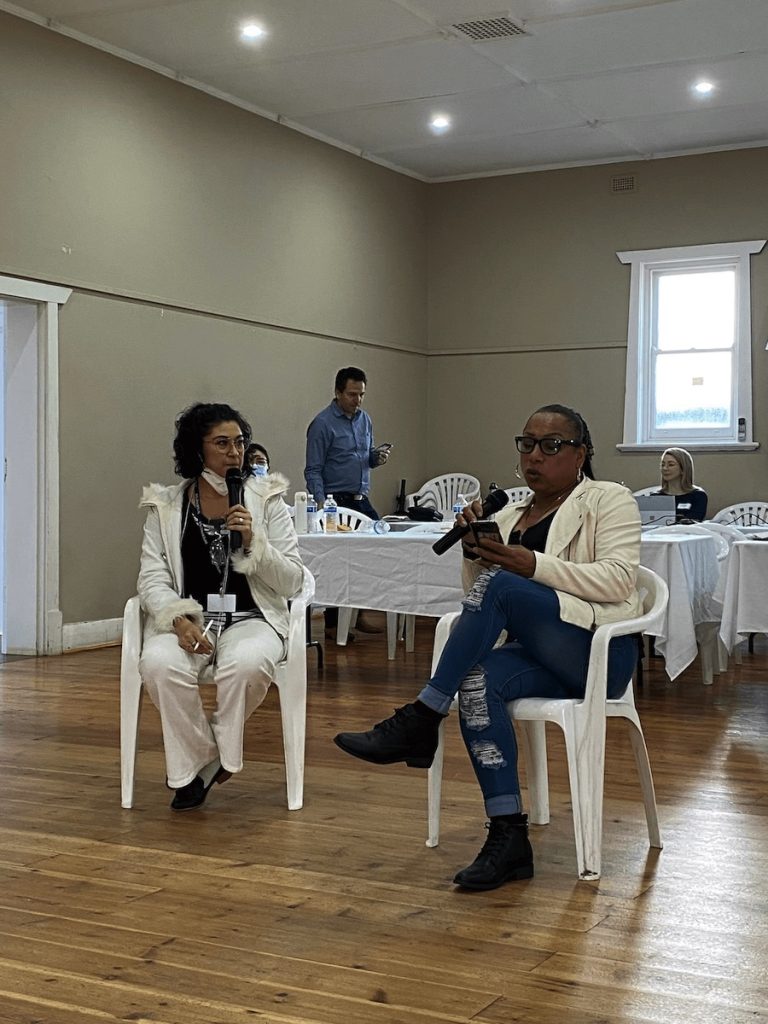 Two people seated on chairs. Alexandra (left) holds the microphone and reads her story.