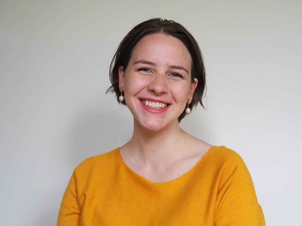 Photo of Tessa smiling at camera. She has short, brown hair as is wearing an orange blouse, and has a light grey background behind her.