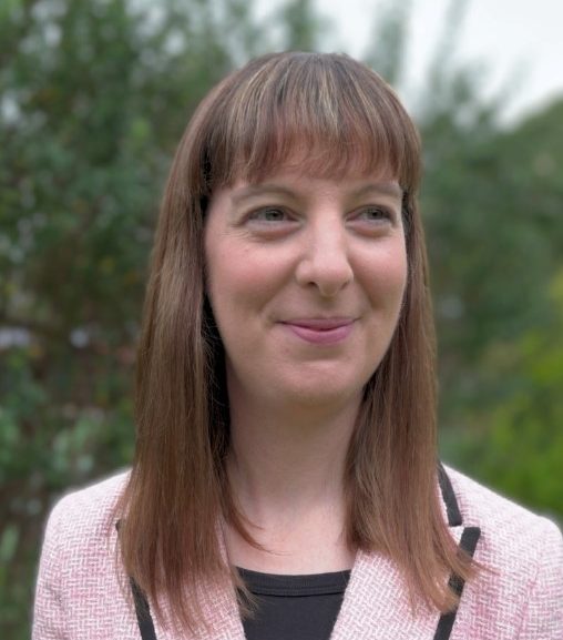 A woman with straight, shoulder-length brown hair and fringe smiles softly while looking slightly to the side. She is wearing a light pink jacket with black trim over a black top. The background is an outdoor setting with blurred greenery.
