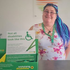 Chealsea stands, smiling at the camera with brown and blue braids in her hair. She stands in front of a sign promoting understanding for hidden disabilities. The text on the sign reads "Not all disabilities look like this" with a graphic of a wheelchair.