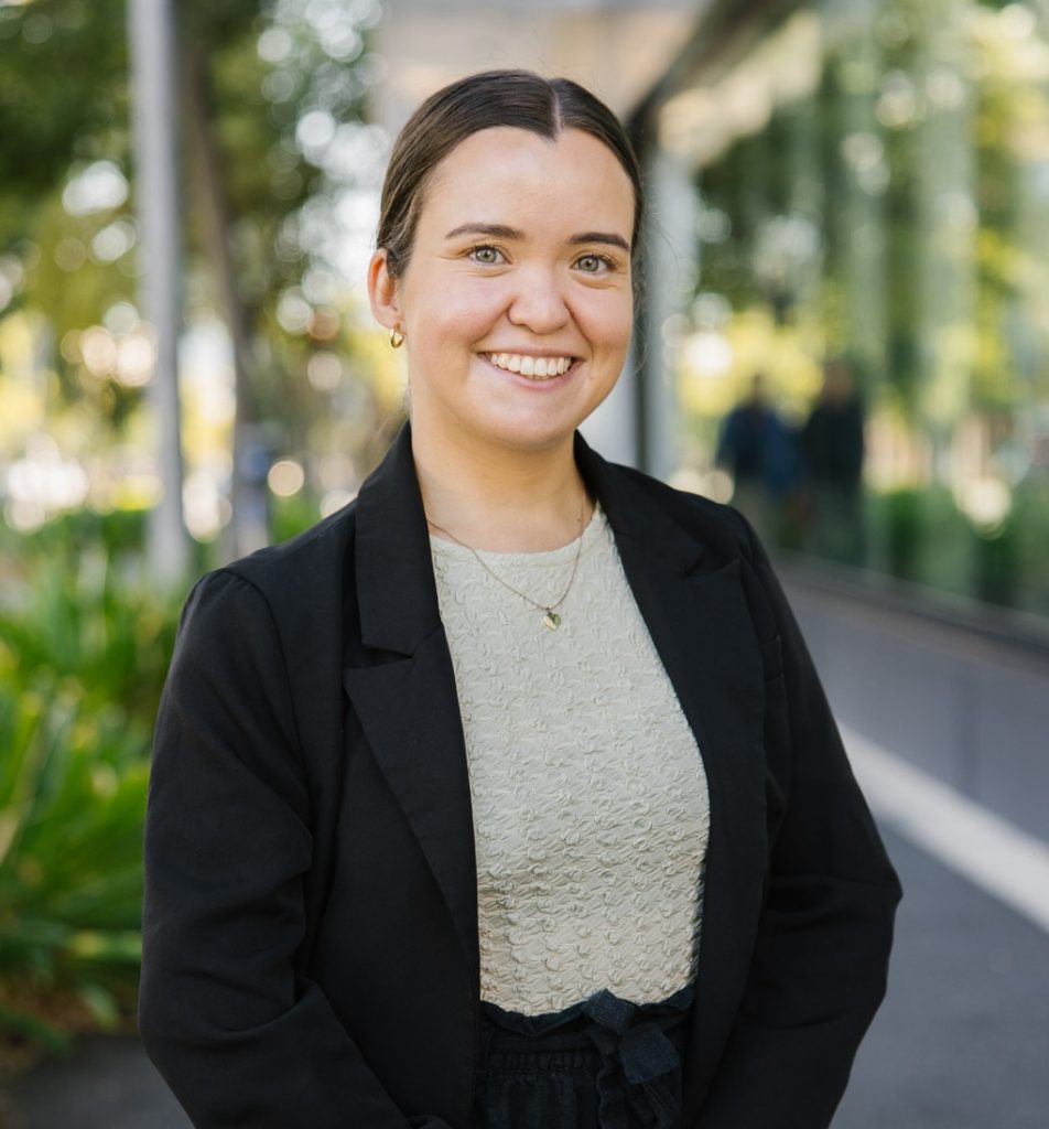 A young woman with dark hair tied back smiles while standing outdoors in a city setting. She is wearing a light textured top, a black blazer, and a gold necklace. Trees and a glass building line the footpath in the background.