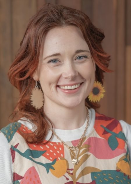 Louie, who has long, layered red hair and sunflower earrings, smiles at the camera. They are wearing a colourful patterned top, in front of a wooden backdrop.