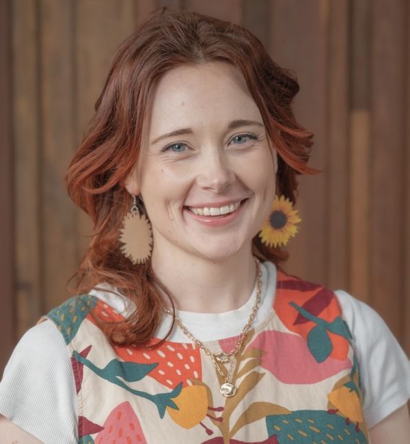 Louie, who has long, layered red hair and sunflower earrings, smiles at the camera. They are wearing a colourful patterned top, in front of a wooden backdrop.