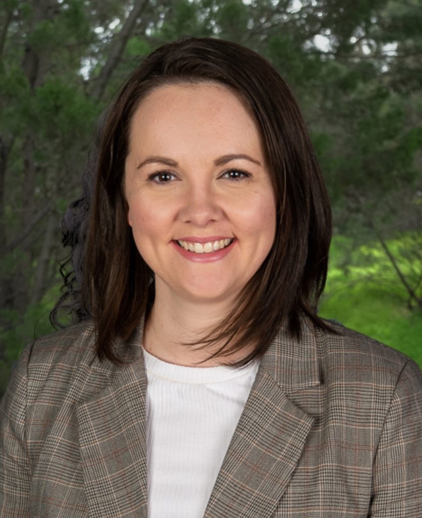 A woman with straight, dark brown hair smiles at the camera while standing outdoors. She is wearing a grey plaid blazer over a white top. The background features softly focused green trees and foliage.