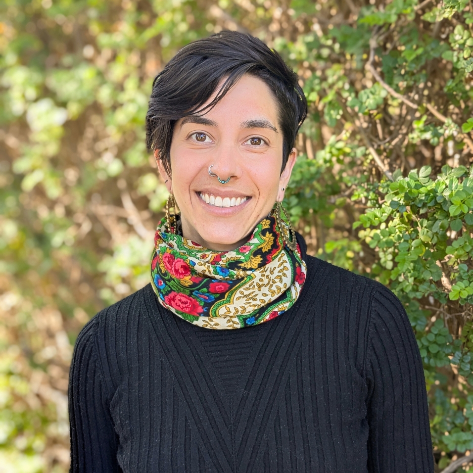 A person with short dark hair smiles warmly while standing in front of a leafy green background. They are wearing a black ribbed top and a colorful patterned scarf with floral and gold designs. They have a septum piercing and are wearing long, decorative earrings.