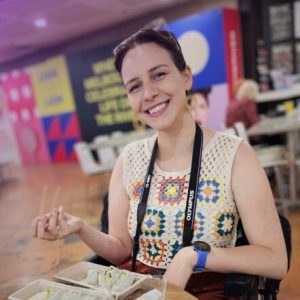 Tessa at a café table, smiling in a sleeveless, crocheted top. She holds chopsticks in one hand.
