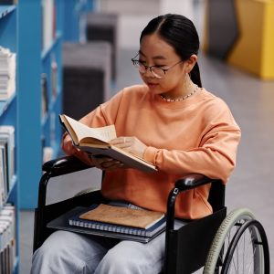 Young female student sits in her wheelchair and reads a textbook next to a blue bookshelf.