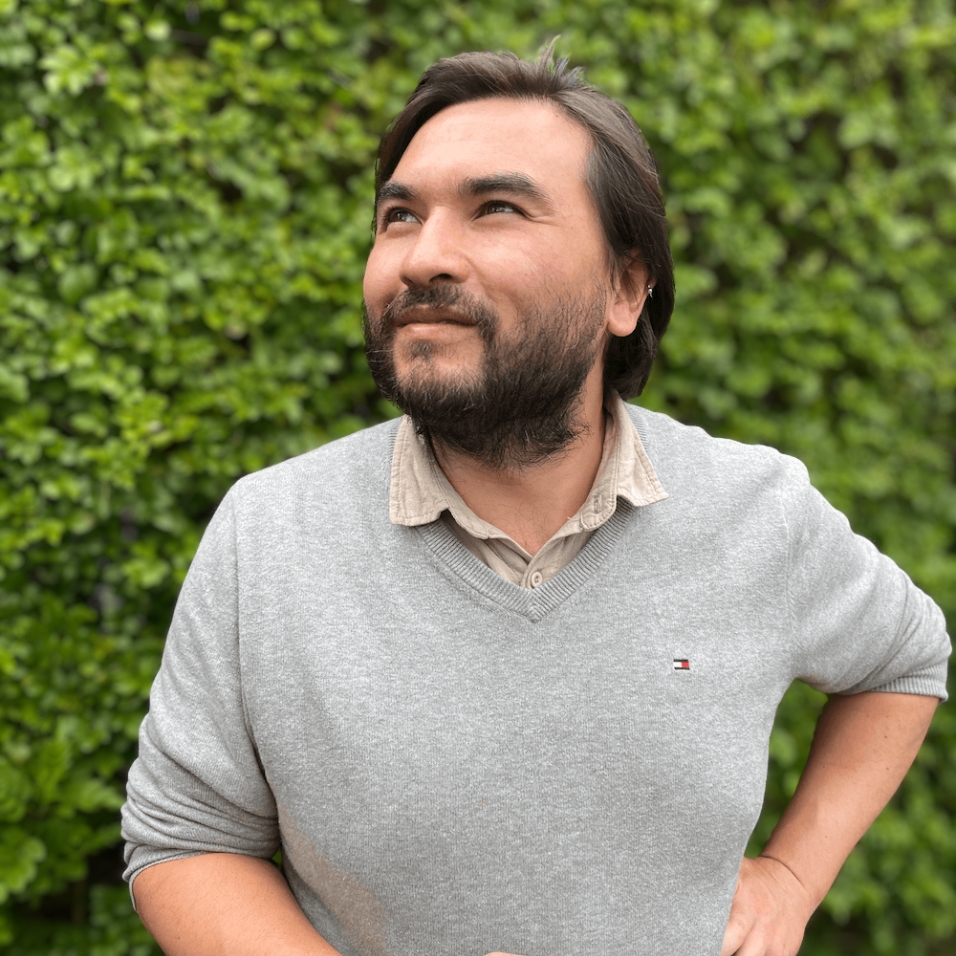 A man with dark hair and a beard looks upward with a thoughtful expression while standing in front of a lush green leafy wall. He is wearing a light grey sweater over a beige collared shirt, with one hand on his hip.