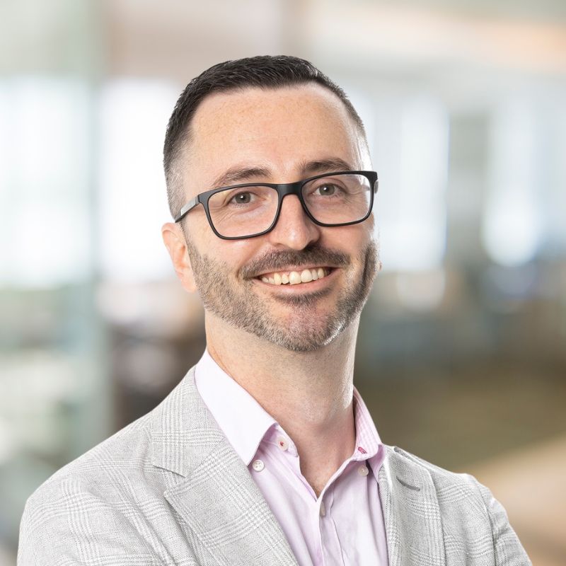A man with short dark hair and glasses smiles confidently at the camera. He is wearing a light grey blazer over a pale pink shirt. The background is a softly blurred modern office space.