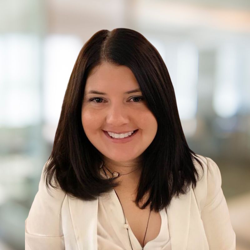 A woman with straight dark brown hair smiles warmly at the camera. She is wearing a white blazer and a light top, with a blurred professional indoor setting in the background.