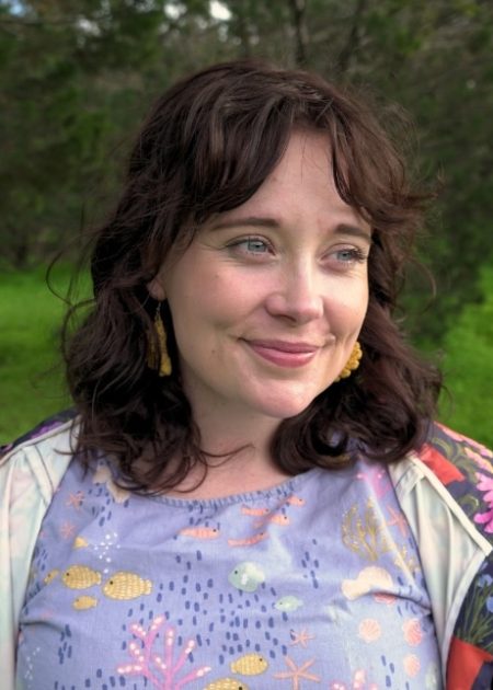 A person with wavy brown hair smiles gently while looking off to the side. They are wearing gold earrings, a lavender top with a colorful underwater-themed pattern, and a floral jacket. The background is blurred greenery.