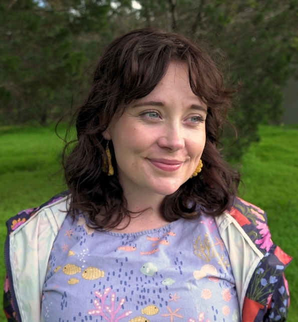A person with wavy brown hair smiles gently while looking off to the side. They are wearing gold earrings, a lavender top with a colorful underwater-themed pattern, and a floral jacket. The background is blurred greenery.
