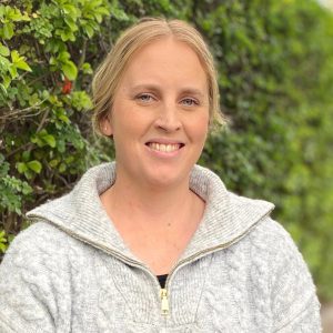A blonde woman in a chunky light-grey cable-knit sweater smiles beside a leafy green hedge.