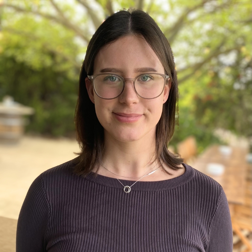 A woman with shoulder-length dark brown hair and clear-framed glasses smiles softly at the camera. She is wearing a ribbed, dark long-sleeve top and a silver necklace with a circular pendant. The background features a blurred outdoor setting with greenery and a long wooden table.