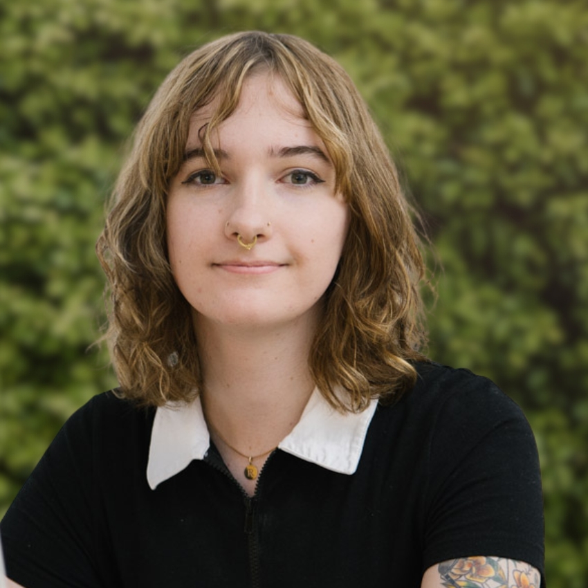 A person with wavy light-brown hair and a septum piercing looks at the camera with a slight smile. She wears a black top with a white collar and a small pendant necklace, with a tattoo visible on one arm. The background is a leafy green hedge in soft focus.