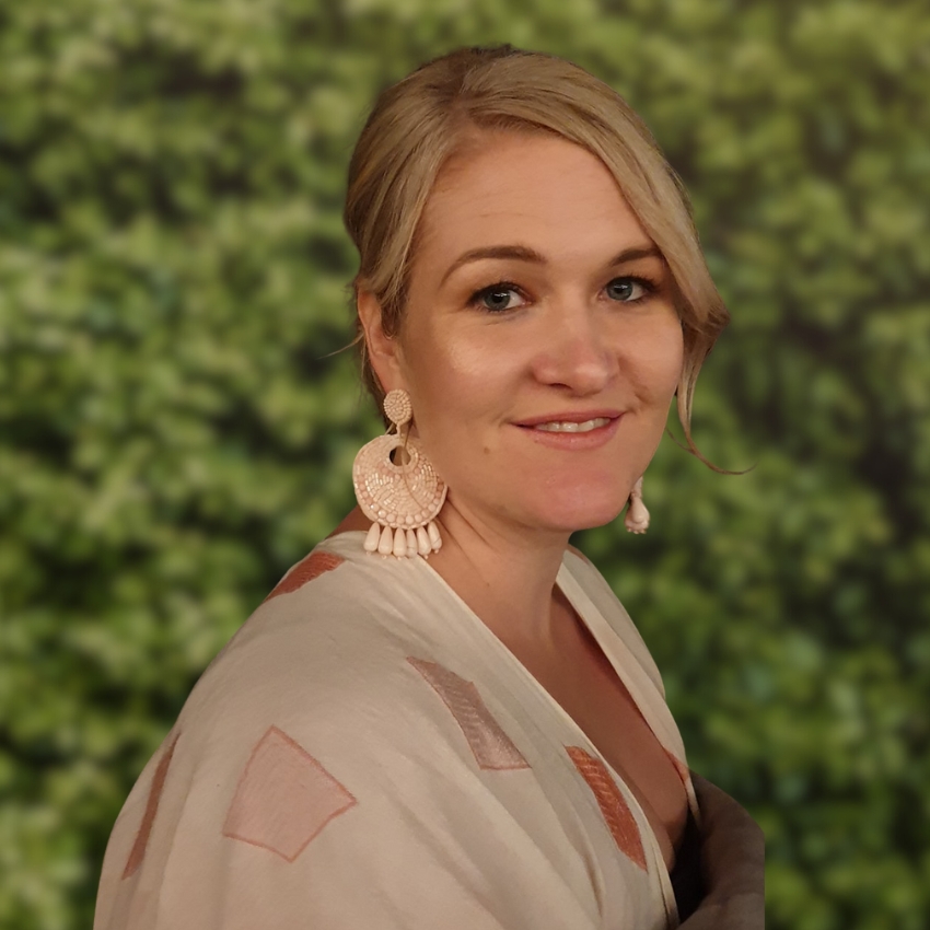 A woman with blonde hair styled to the side smiles softly at the camera. She wears large pale pink earrings and a light patterned top. The background is a softly blurred wall of green foliage.