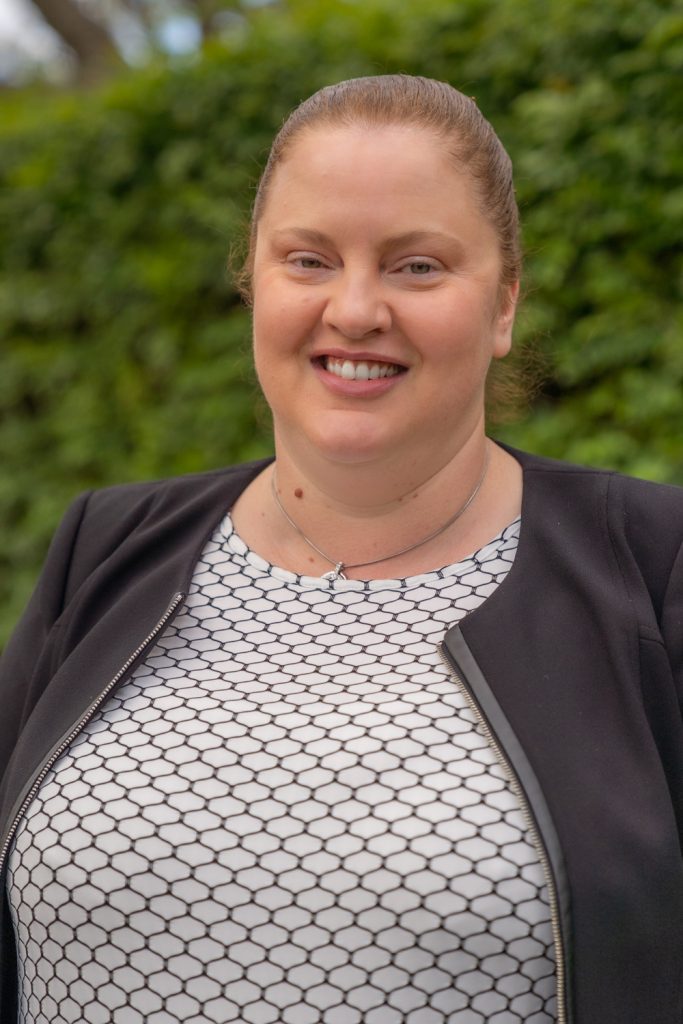 A person with light brown hair in a ponytail, wearing a geometric patterned white and black shirt and a black jacket. They are smiling at the camera.